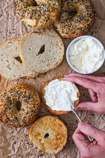 A person spreading cream cheese over the surface of a halved homemade everything bagel on a piece of brown paper next to other bagels and a bowl of cream cheese.