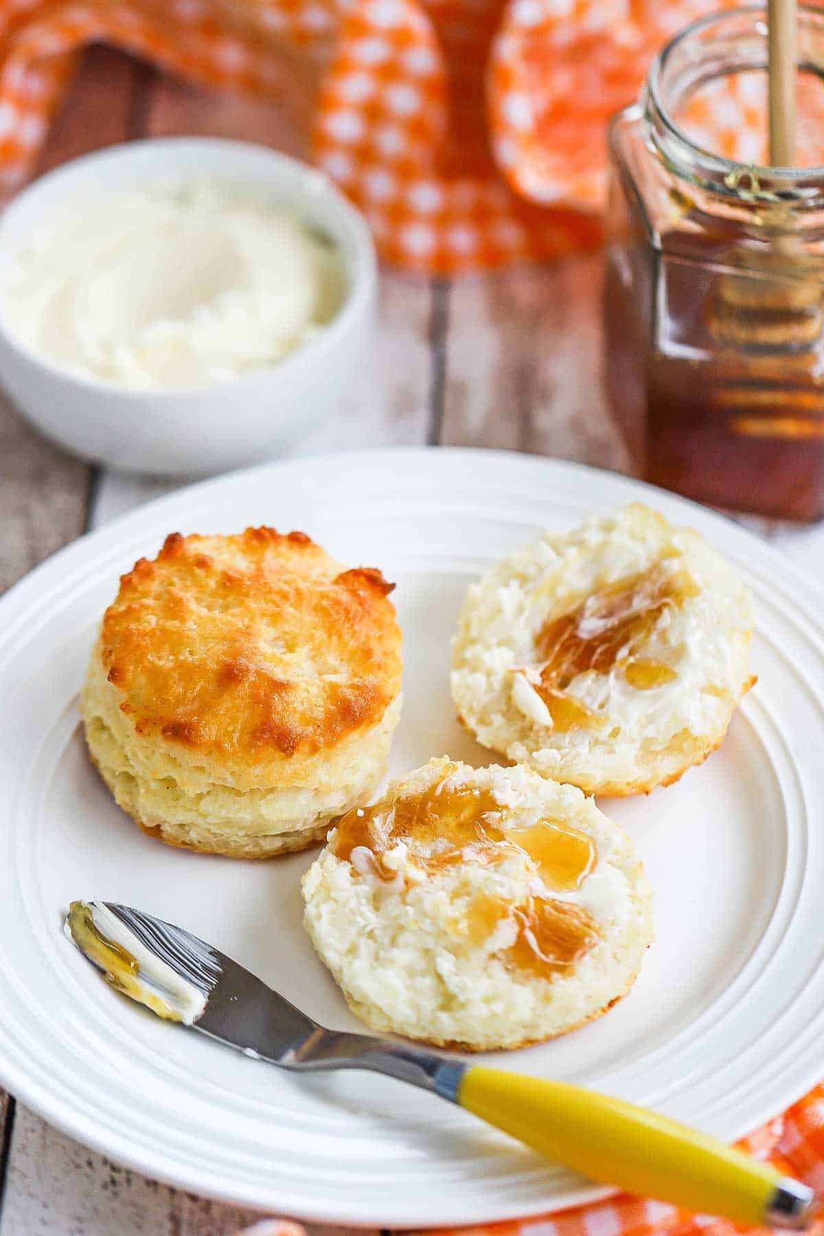 A small white plate filled with two buttermilk biscuits, one that has been split in half and has butter and honey spread on it and the other untouched.