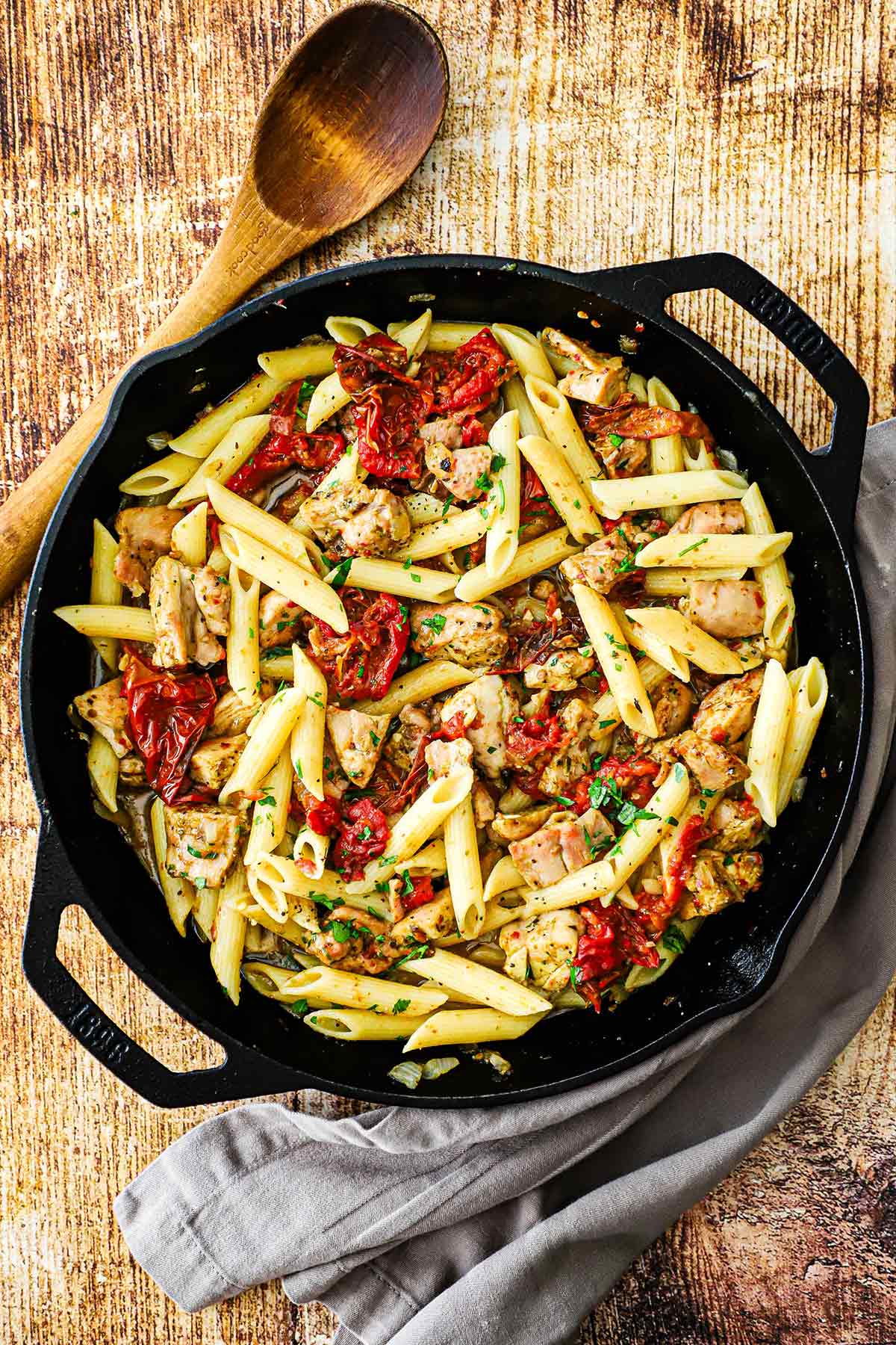 A large black cast-iron skillet filled with pasta with roasted tomatoes and pesto chicken and a large wooden spoon next to it.