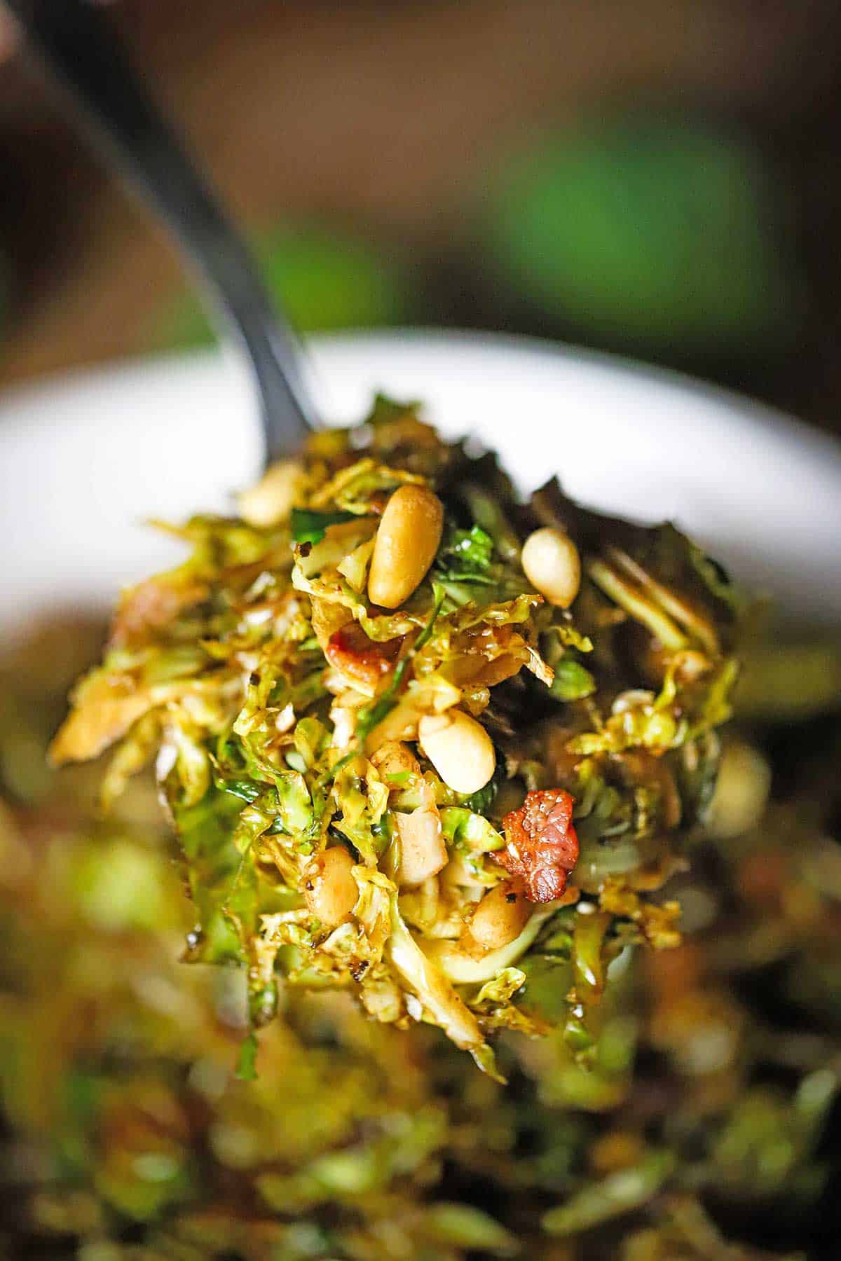 A large serving spoon being lifted holding a serving of shaved Brussles sprouts with balsamic and pancetta over a bowl of the same.