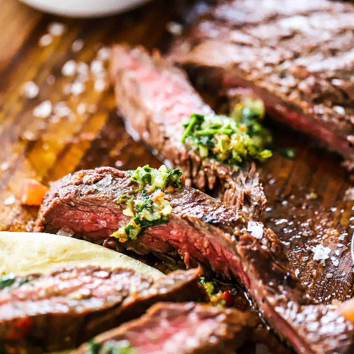 Strips of medium-rare grilled carne asada on a wooden cutting board with small amounts of chimichurri sauce on top as well as a sprinkling of coarse sea salt.