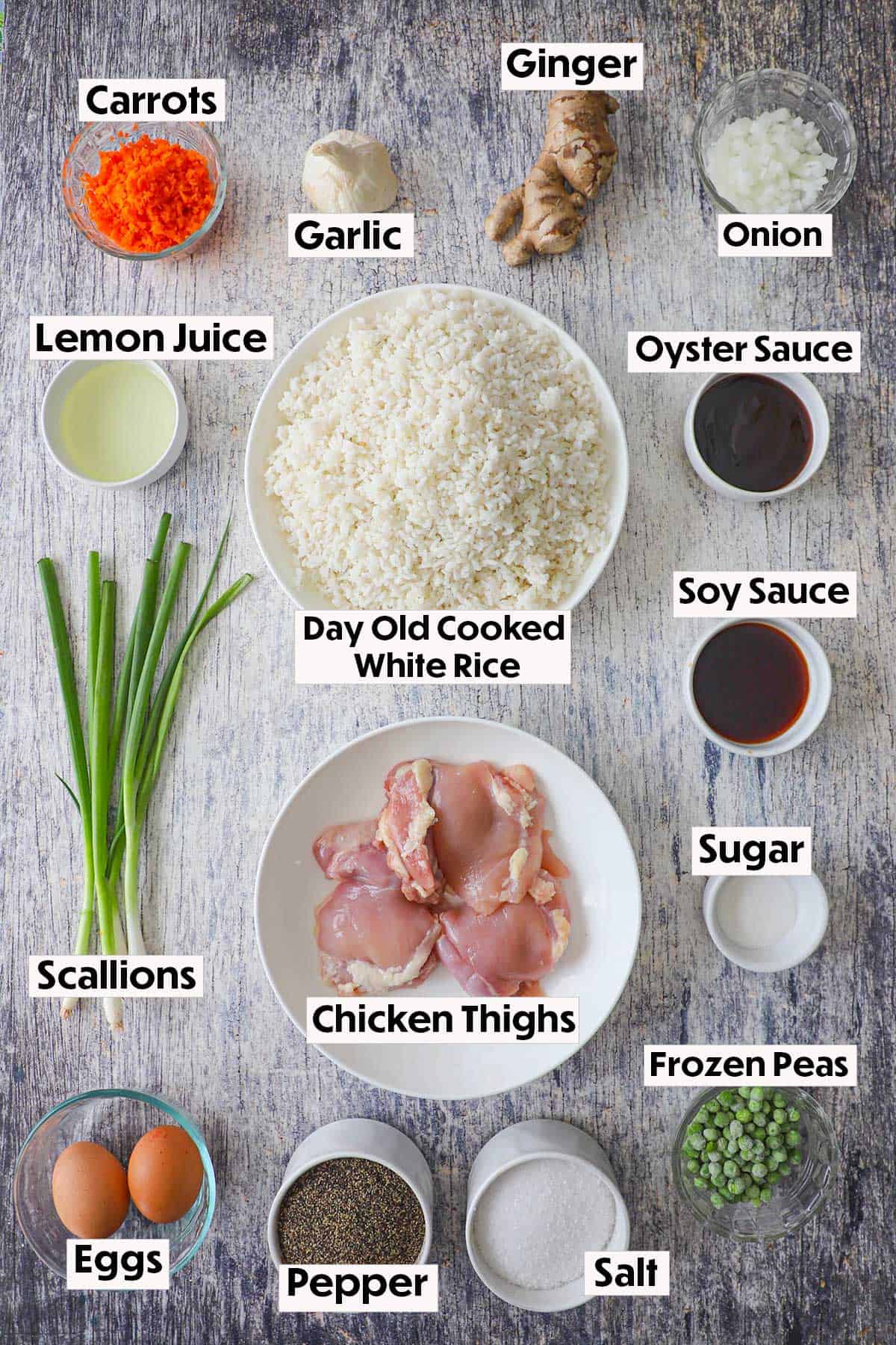 An arrangement of ingredients on a grey wooden background including rice, chicken, carrots, scallions, onion, eggs, peas, ginger, garlic, and seasonings.