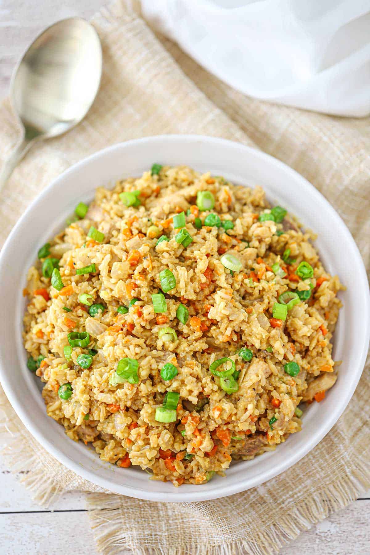 A shallow white ceramic bowl filled with a serving of chicken fried rice topped with chopped scallions and a serving spoon nearby.