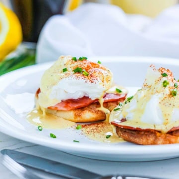 A white plate filled with two Eggs Benedict with toasted English muffins, Canadian bacon, poached eggs, Hollandaise sauce, and garnished with paprika and snipped chives.