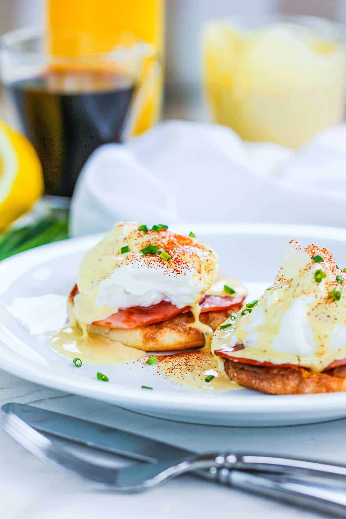 A serving of eggs Benedict on a white plate with a glass cup of coffee and a carafe of orange juice in the background.