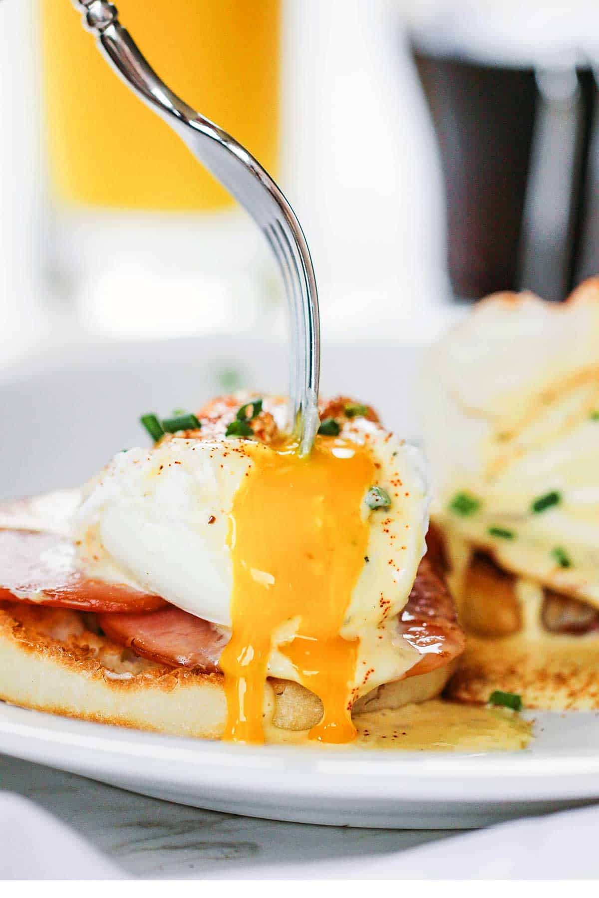 A person using a fork to pierce open a poached egg with a runny yolk spilling over the sides of an Eggs Benedict.