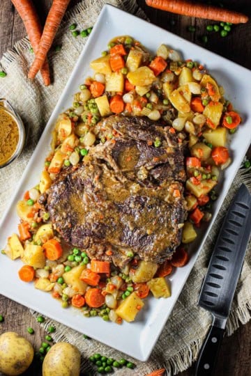 A fully cooked classic pot roast on a large rectangular white platter surrounded by cooked potatoes, carrots, and peas.