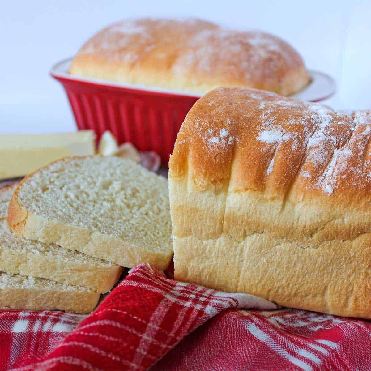 A loaf of country white bread that has several slices cut from it and then another loaf in a burgundy ceramic loaf pan with a baked loaf in the background.