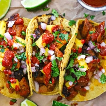 Three grilled tacos al pastor pressed against each other loaded with marinated grilled pork, pineapple chunks, cilantro, red onion, and a homemade sauce on top.