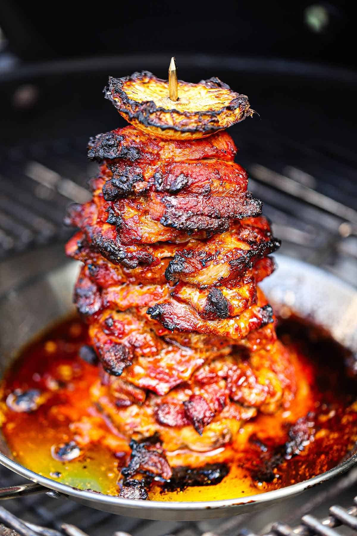 A tower of grilled layers of marinated pork with a rod centered in the middle anchoring the meat with a slice of pineapple on top all on a charcoal grill.