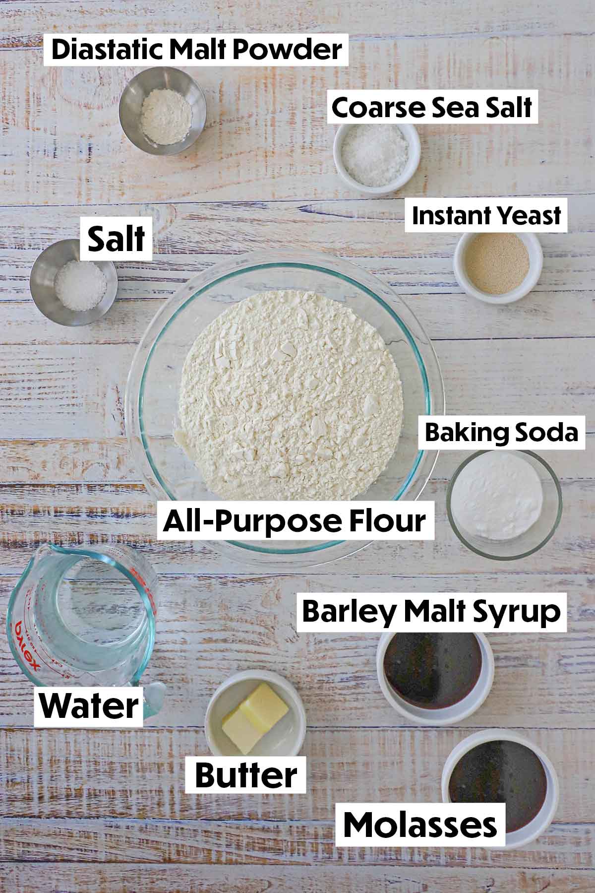 An arrangement of ingredients for homemade pretzel buns on a white background including flour, yeast, salt, malt powder, baking soda, water, butter, and molasses.
