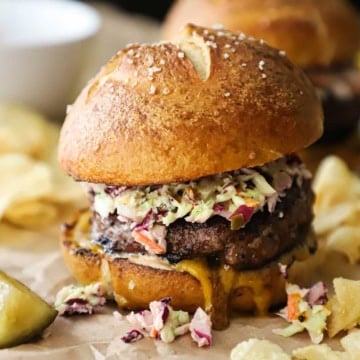 A pork burger topped with creamy coleslaw on a homemade pretzel bun with Carolina mustard sauce oozing from the underneath the patty with kettle chips scattered around the burger.