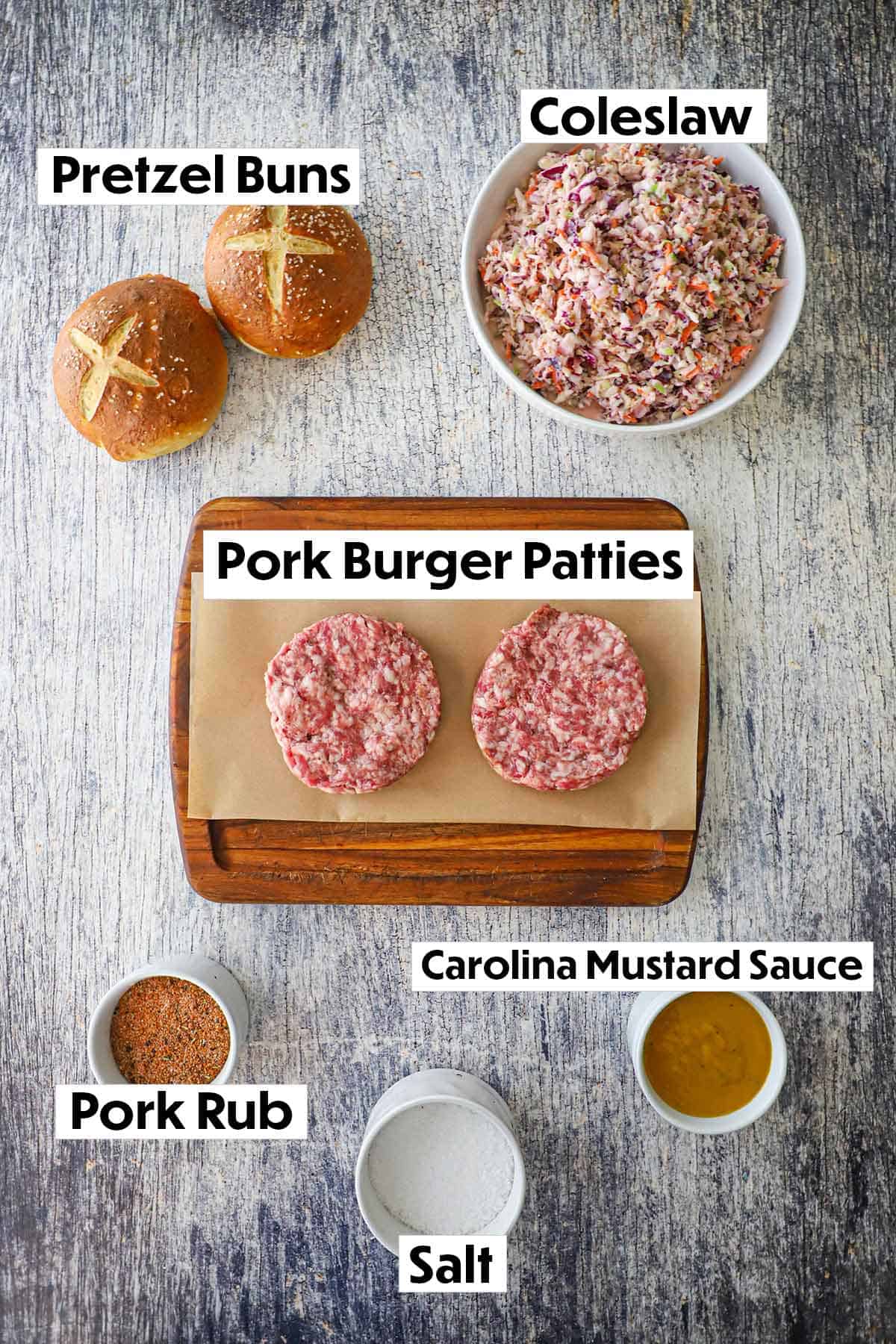 An arrangement of ingredients for pork burgers with pretzel buns on a grey wooden background including pretzel buns, coleslaw, pork burger patties, pork rub, salt, and mustard sauce.