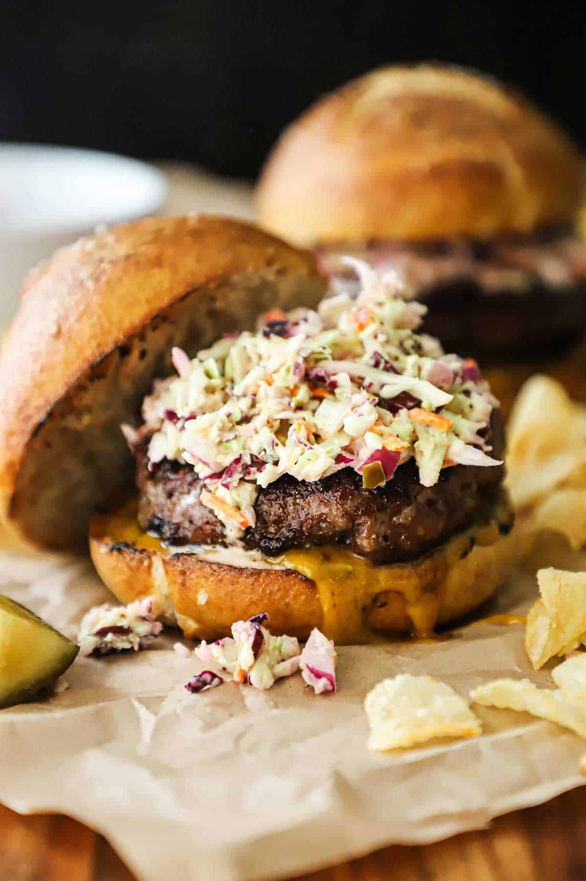 Two pork burgers with pretzel buns sitting on crumpled brown paper with kettle chips scattered around.