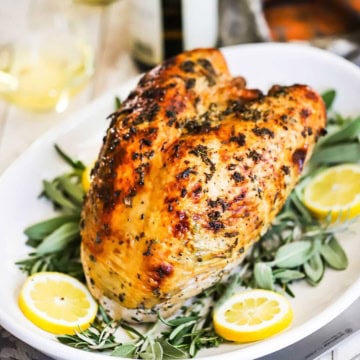 A Roast Turkey Breast with Herbs resting on a white platter that is garnished with fresh sage leaves and lemon slices.