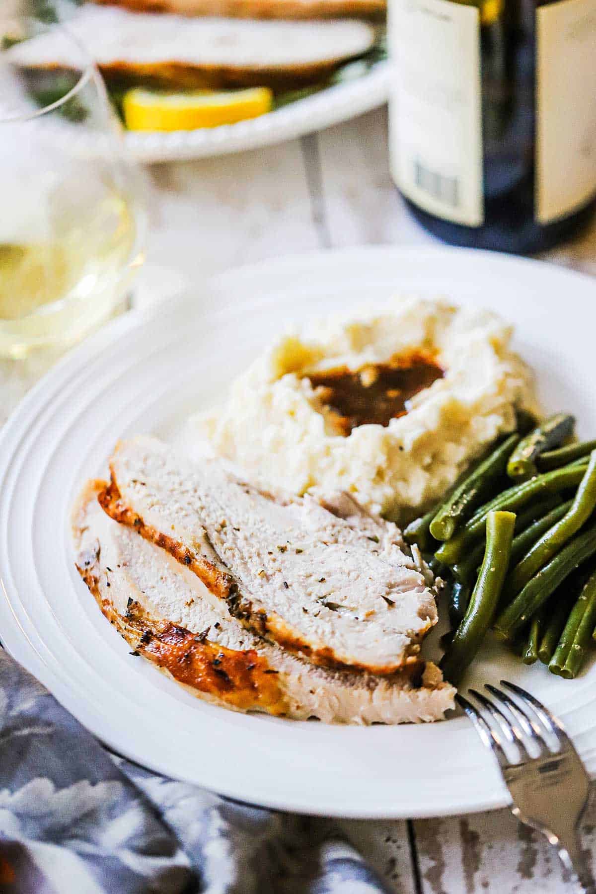 A white dinner plate filled with two slices of roast turkey breast with herbs and a pile of simmered green beans and mashed potatoes topped with gravy.