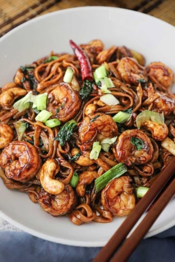 A white bowl filled with shrimp lo mein with cashews garnished with chopped scallions on top and two brown chopsticks resting side by side on the edge of the bowl.