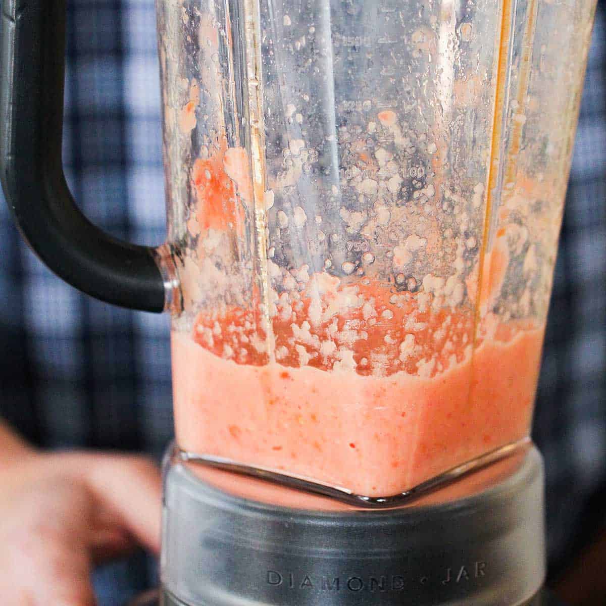 A person standing behind a blender filled with puréed tomatoes, onions, and spices.
