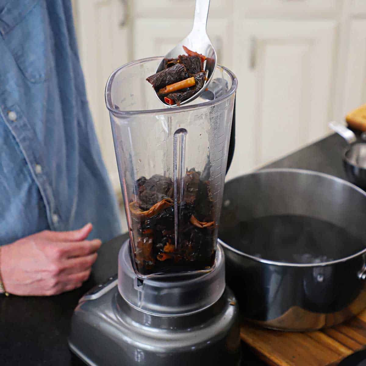 A person using a slotted spoon to transfer freshly boiled dried chili peppers from a pot of hot water into a blender filled with more softened chiles.