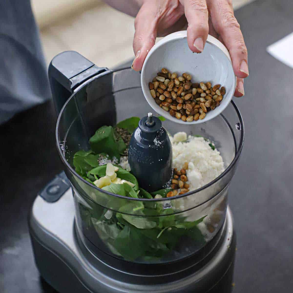 A person transferring toasted pine nuts from a small white bowl into the bowl of a food-processor that is filled with fresh basil, garlic, and grated Parmesan cheese.
