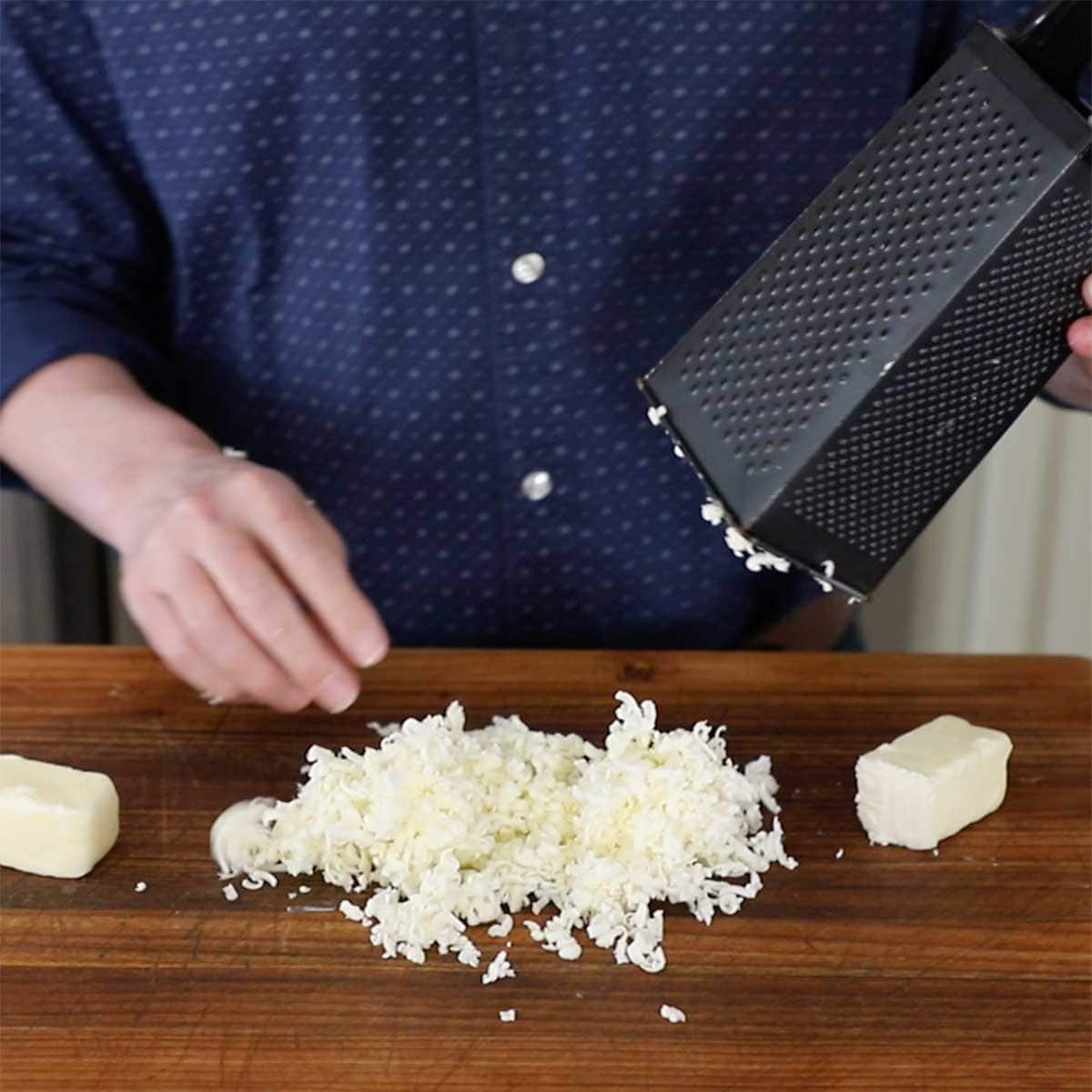 A person raising a black box grater off of a pile of freshly grated frozen butter on a wooden cutting board.