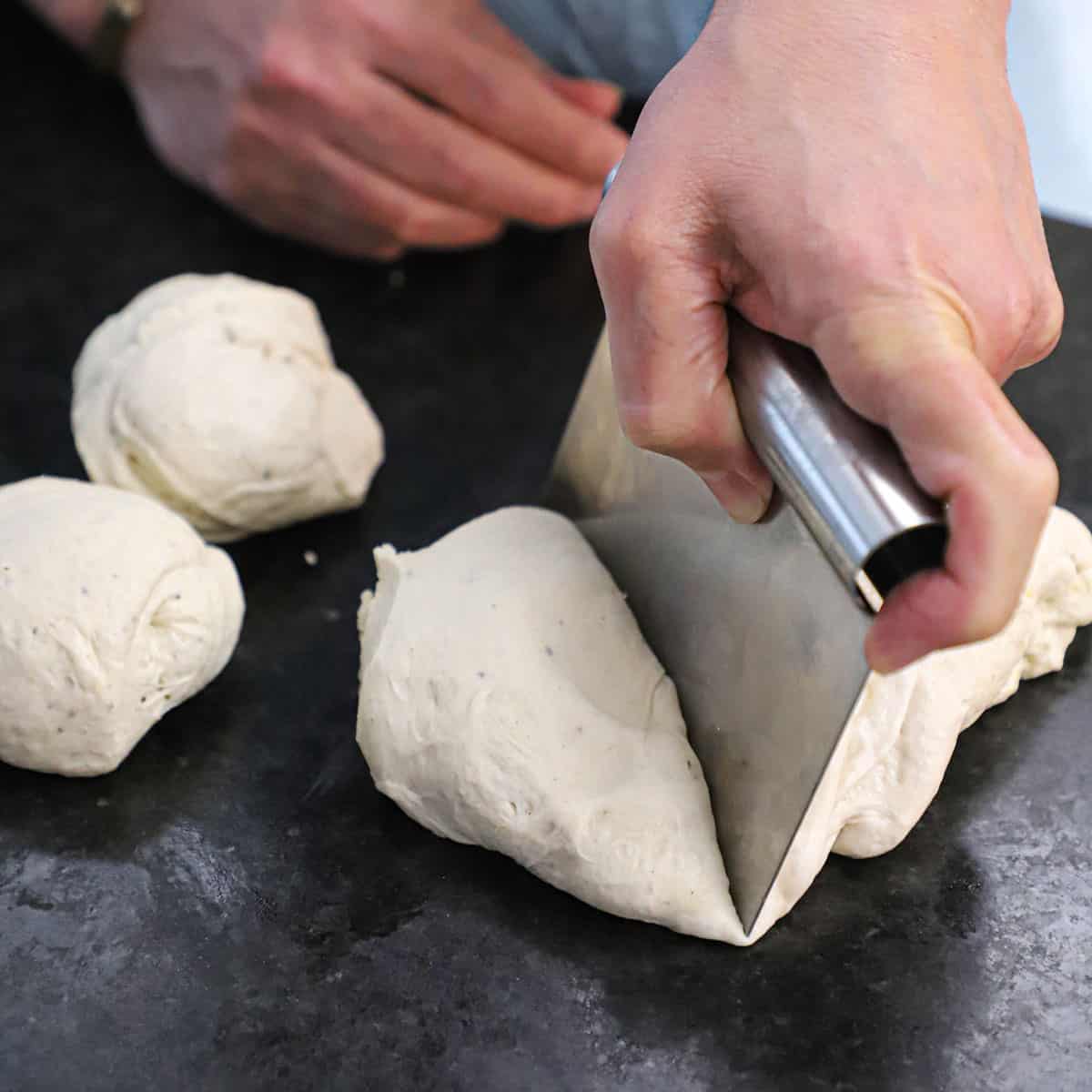 A person using a bench scraper to divide bagel dough into 5 separate pieces on a black marble counter.