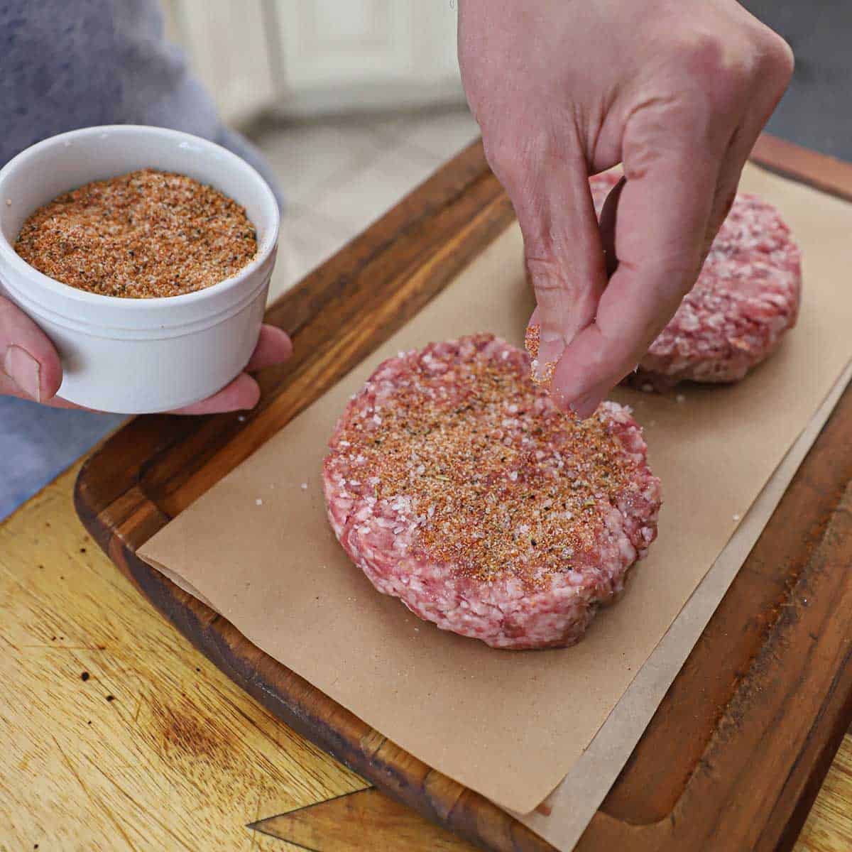 A person using his hand to sprinkle pork rub over the top of an uncooked pork burger patty on wax paper on a small cutting board.