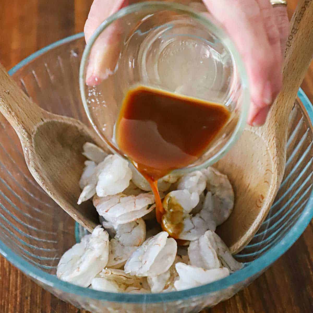 A person pouring a soy sauce and Chinese wine marinade over uncooked shrimp in a small glass bowl.