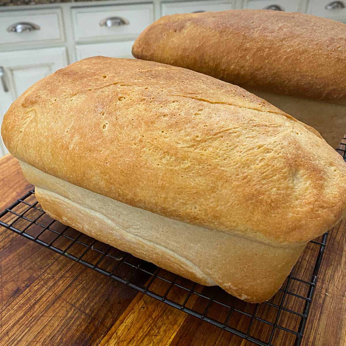 Two large unsliced loaves of country white bread cooling on a baking rack on top of a wooden cutting board.