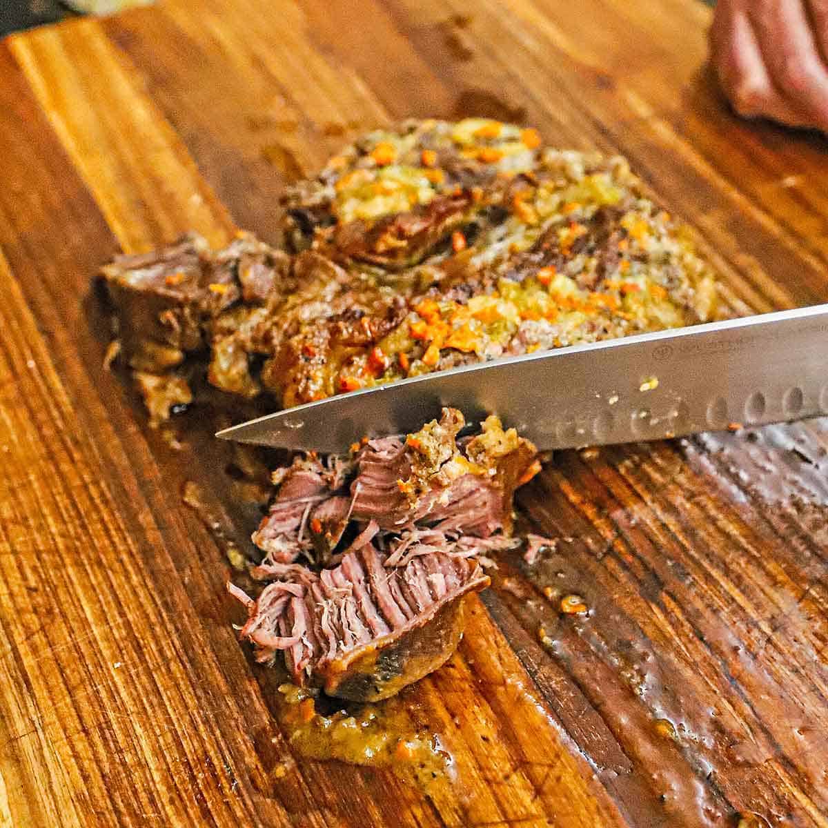 A person using a large chef's knife to cut a classic pot roast into bite-sized pieces on a large wooden cutting board.
