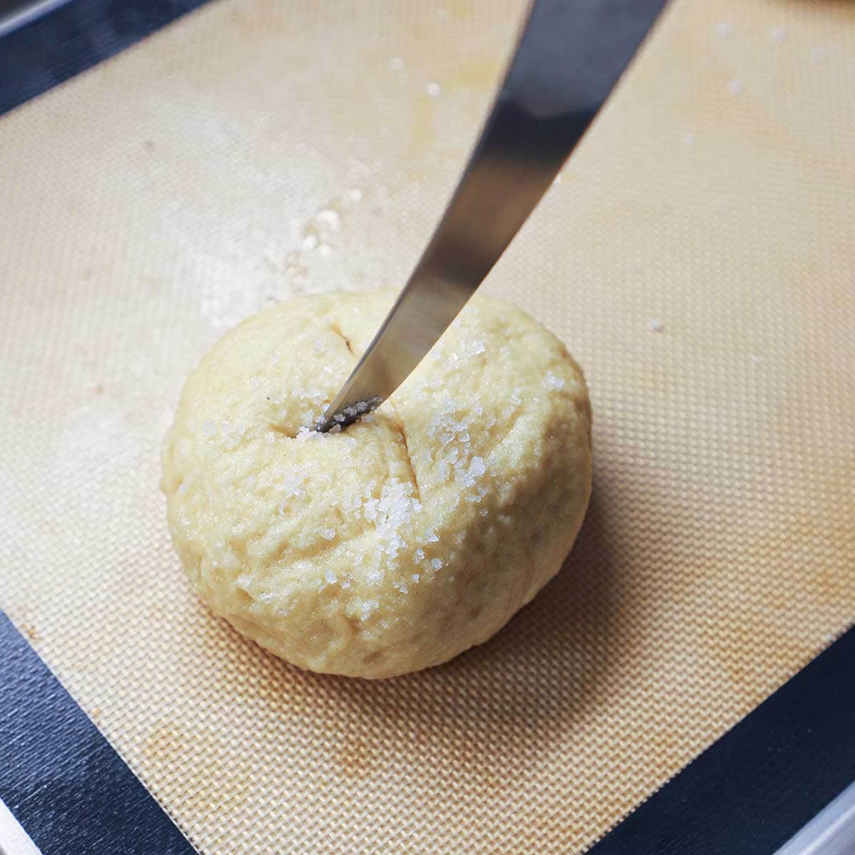 A person using a sharp knife to cut an 'x' in the top of a partially cooked pretzel bun resting on a silicone matt in a baking sheet.