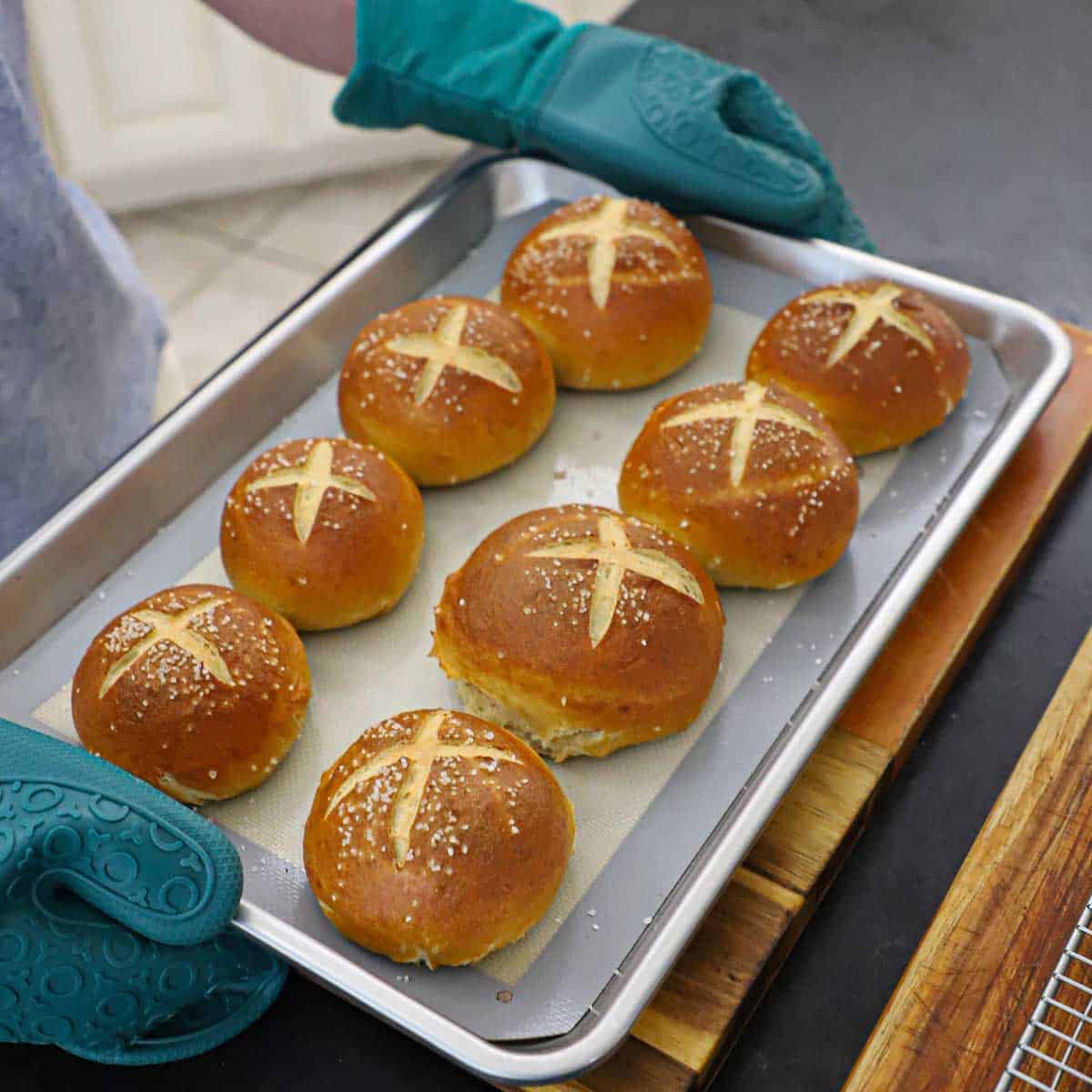 A person holding a baking sheet pan that is filled with eight freshly baked homemade pretzel buns.