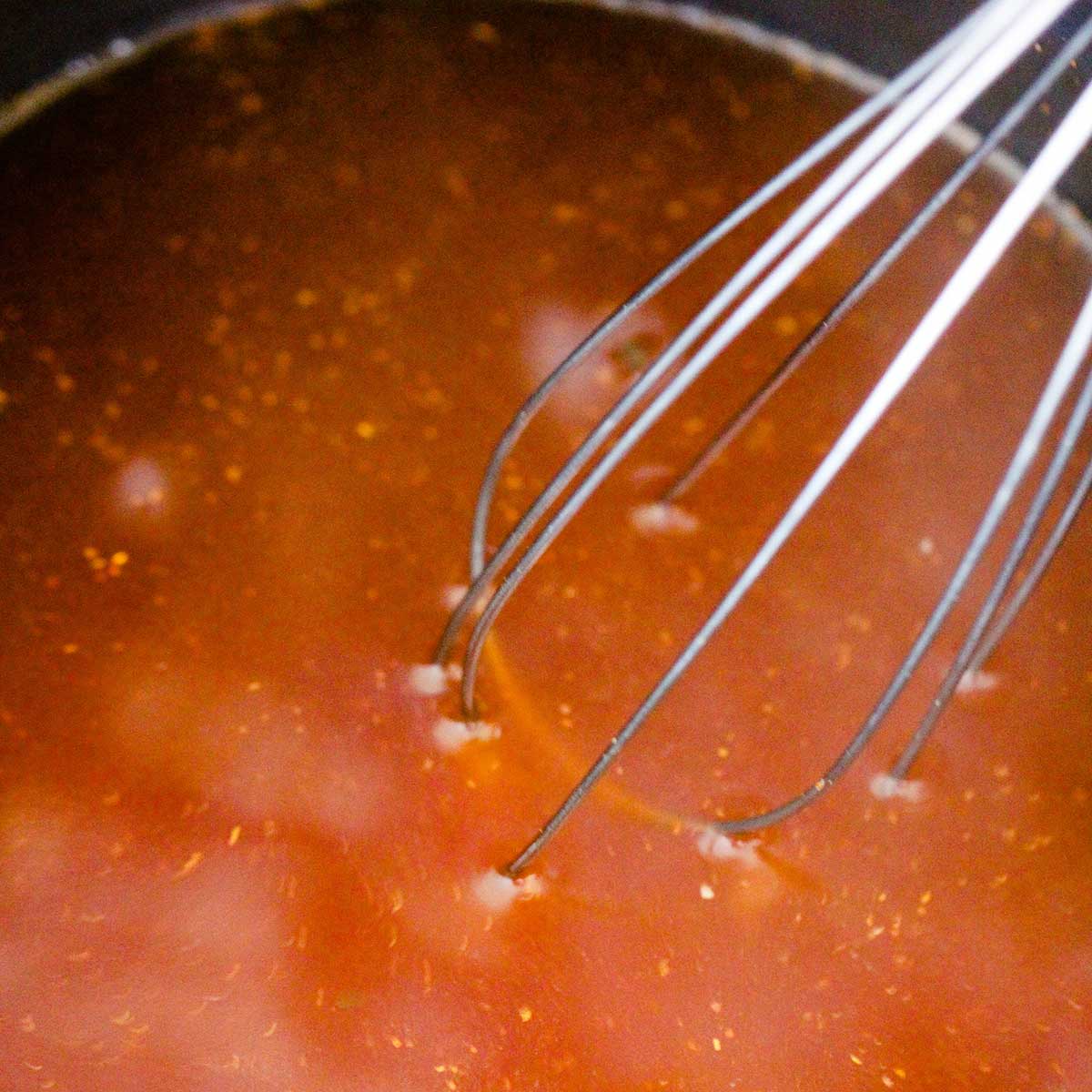 A whisk being used to whisk chicken stock in a pan that has chili powder and tomato bouillon dissolved into it.