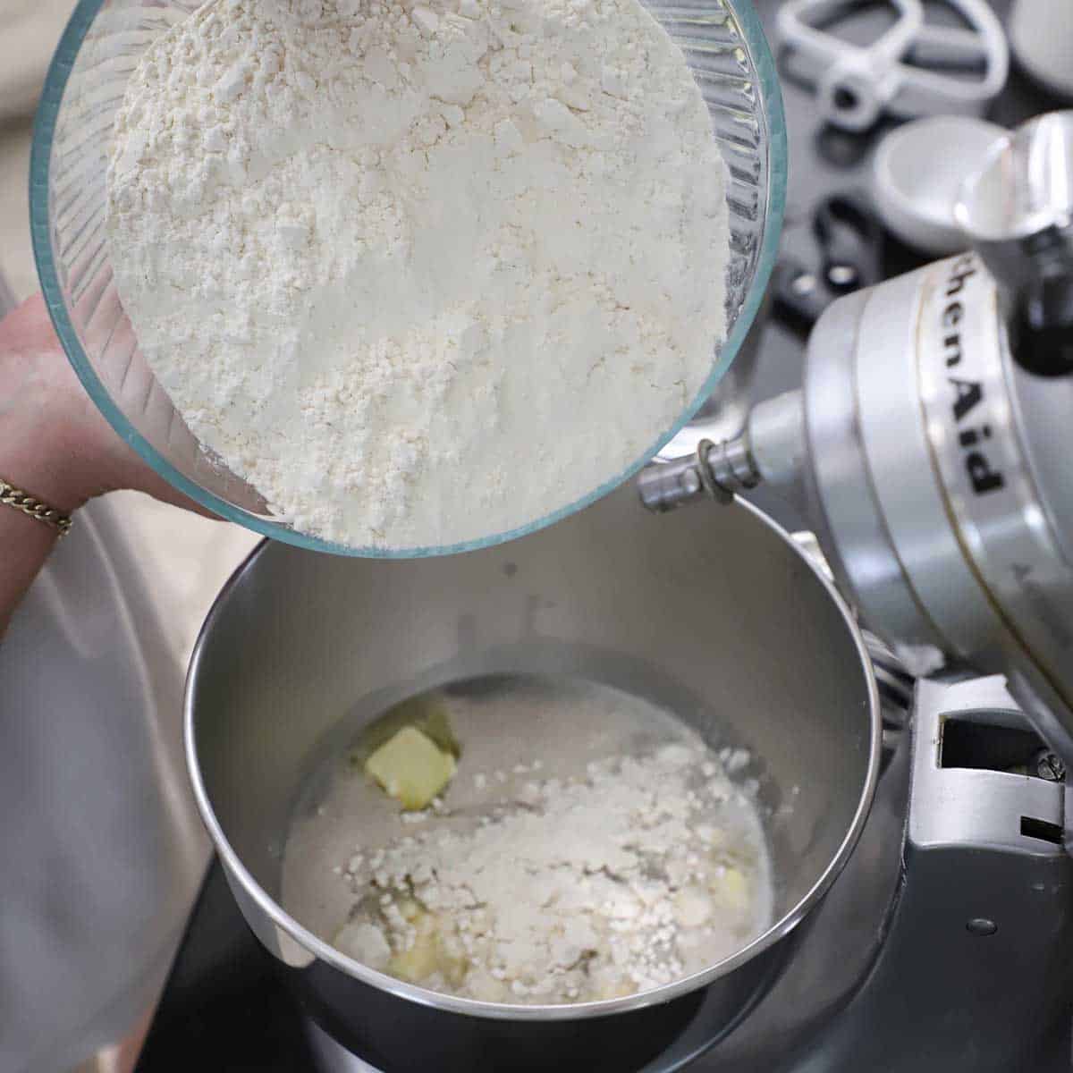 A person adding all-purpose flour from a glass bowl into the bowl of a stand-mixer that is filled with foamy yeast water and cubes of softened butter.