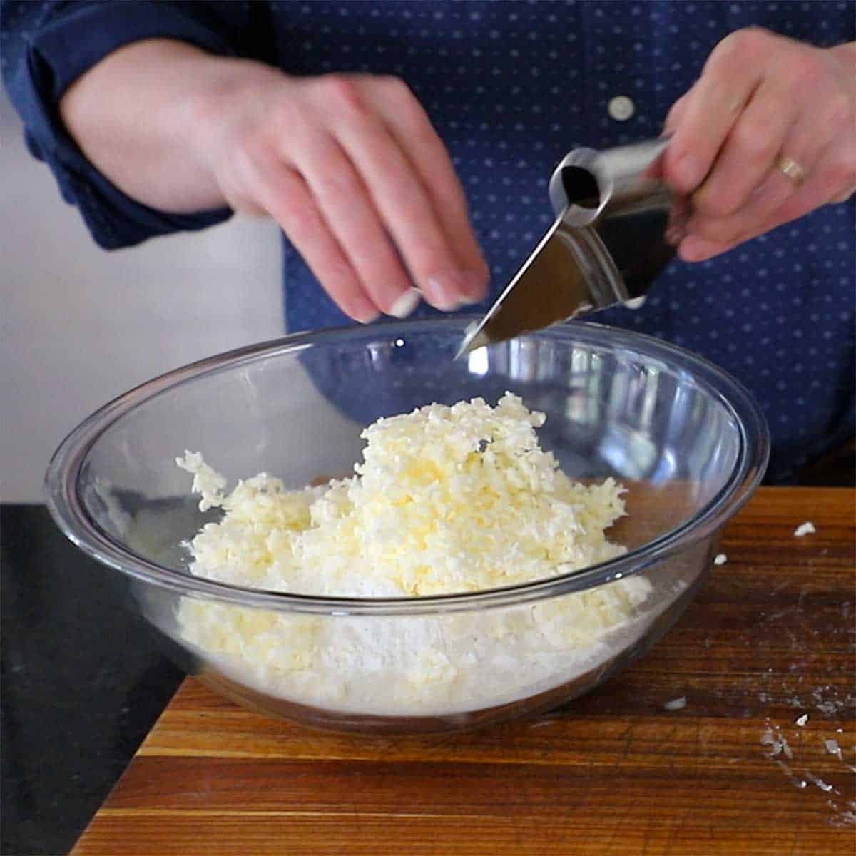A person using a metal bench scraper to transfer grated butter into a glass bowl filled with self-rising flour.