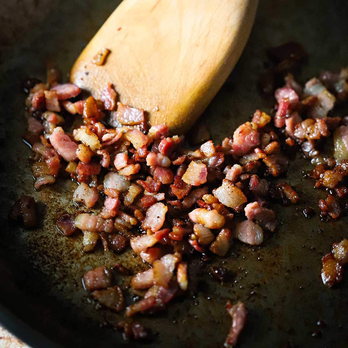 Cubed pancetta being cooked until crispy in a skillet with a large wooden spatula stirring it.