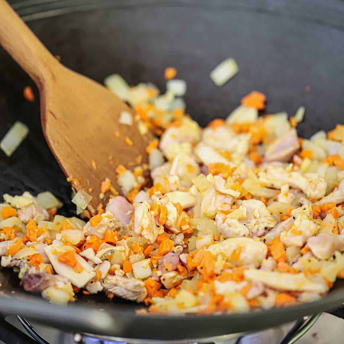 A person using a wooden spatula to stir chunks of chicken meat with sautéd vegetables in a black wok on a gas stove.