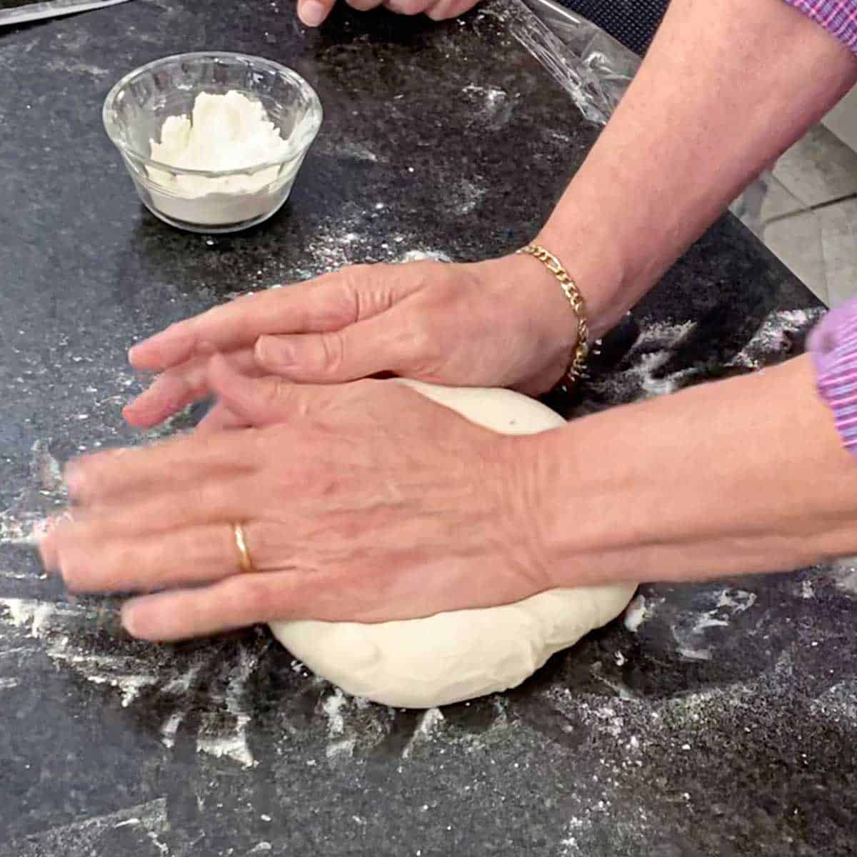 A person kneading bagel dough on a lightly flour black marble counter.