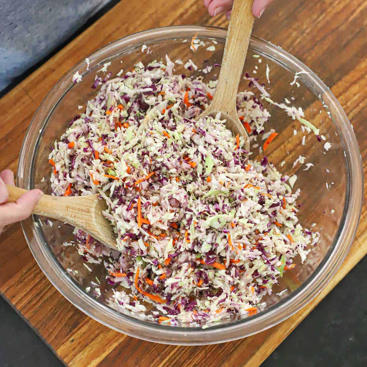 A person using two large wooden spoons to toss together shredded green cabbage, red cabbage, and shredded carrot in a large glass bowl.