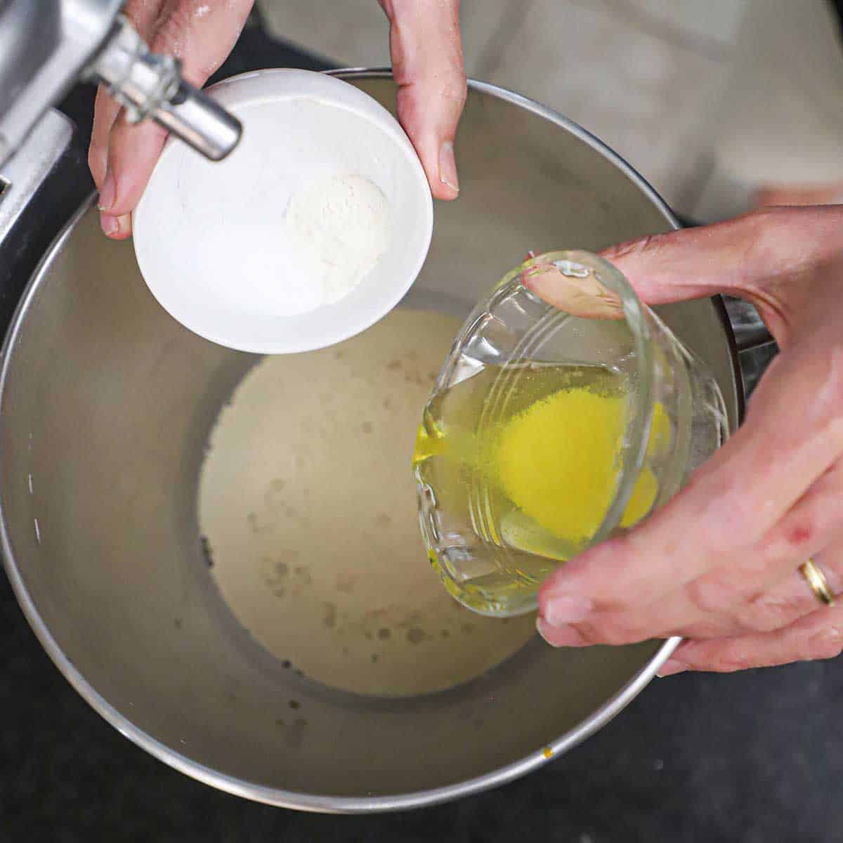 A person dumping baking soda from a small bowl and melted butter from another small bowl into the bowl of a stand mixer that is filled with foamy yeast water.