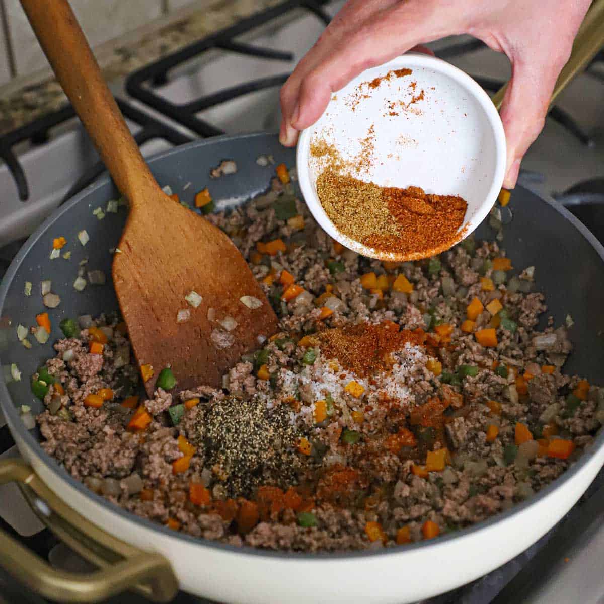 A person dumping chili powder and ground cumin from a small bowl into a skillet filled with cooked ground beef and sautéed chopped onions and peppers.