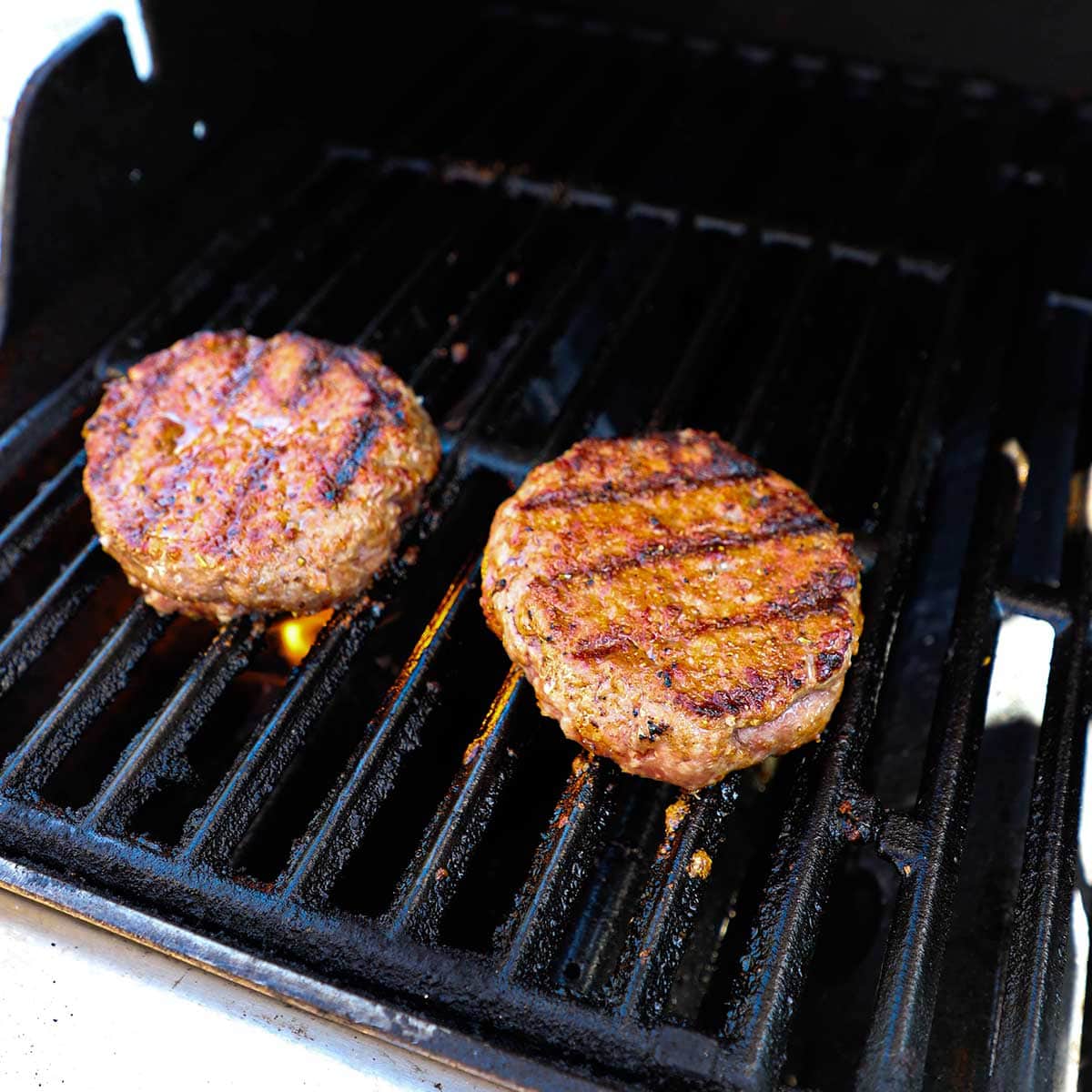 Two pork burgers being grilled on a gas grill with grill marks visible on the top of the burgers.