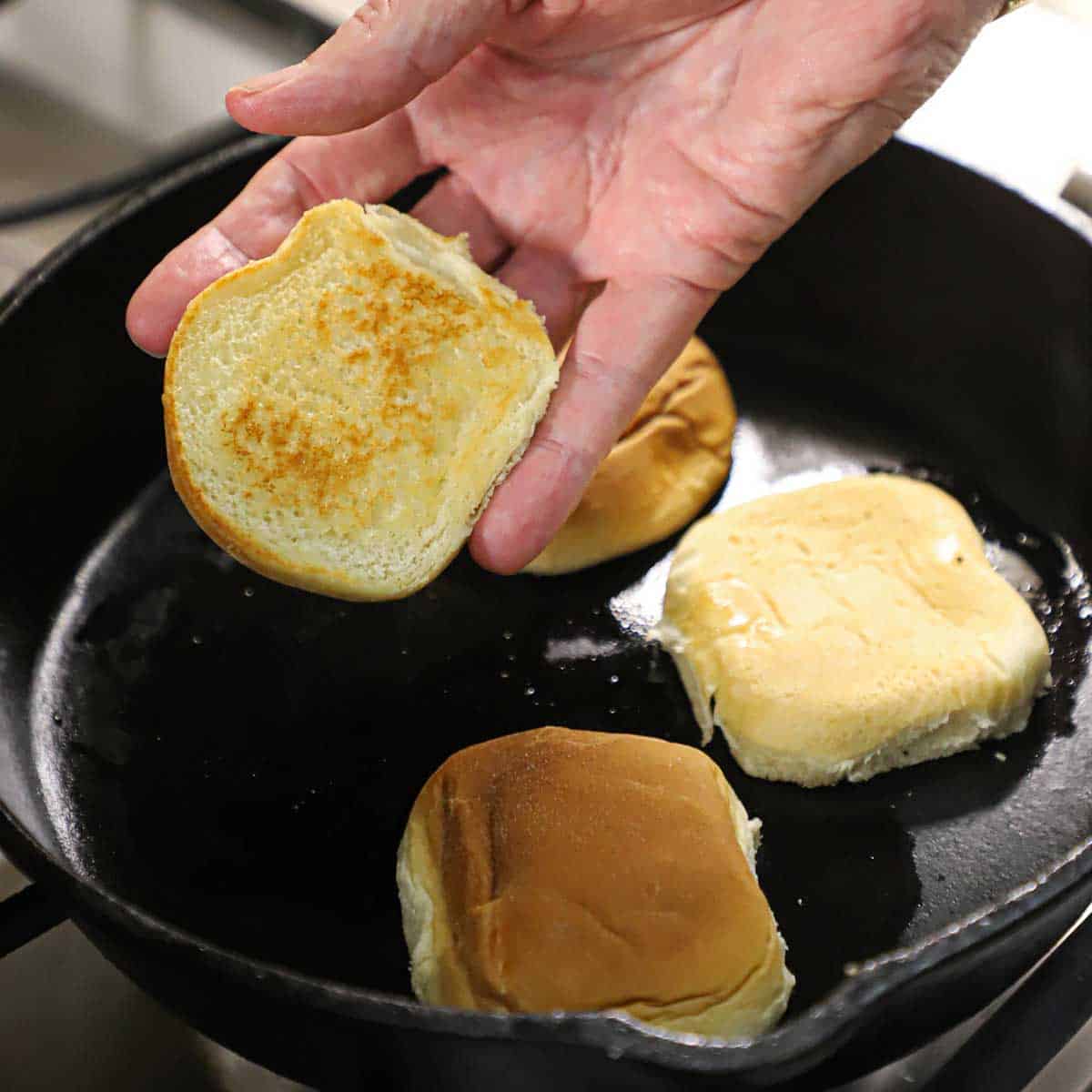 A person lifting up the bottom of a slider bun that has been toasted in a cast-iron skillet with three other slider buns being toasted cut-side down.