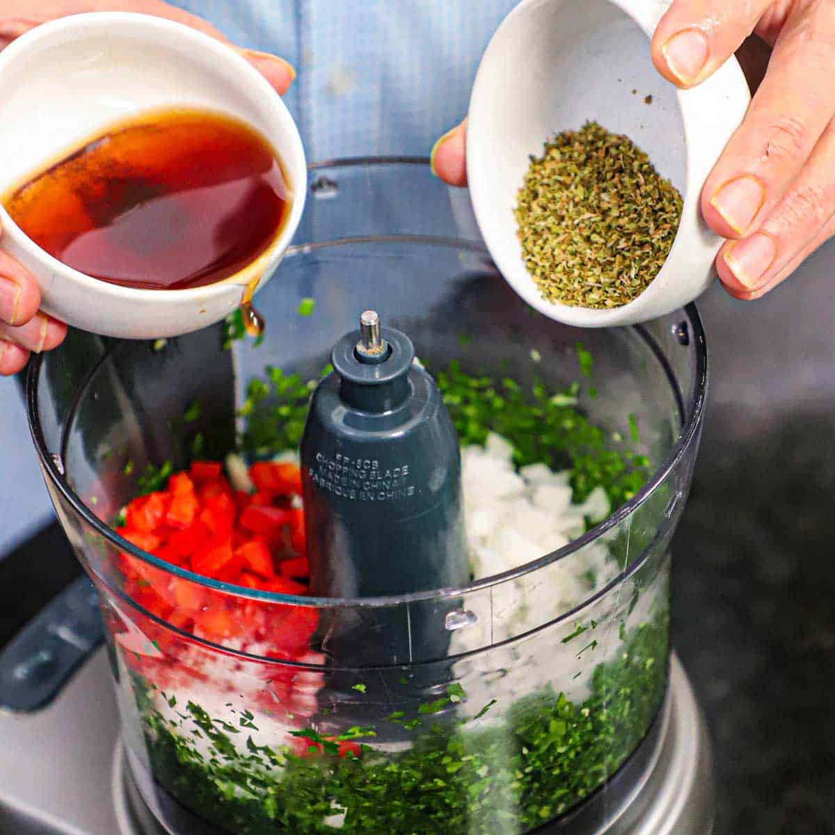 A person using both hands to transfer red wine vinegar from a small bowl and dried oregano from another small bowl into a food processor filled with chopped parsley, onion, and red bell pepper.