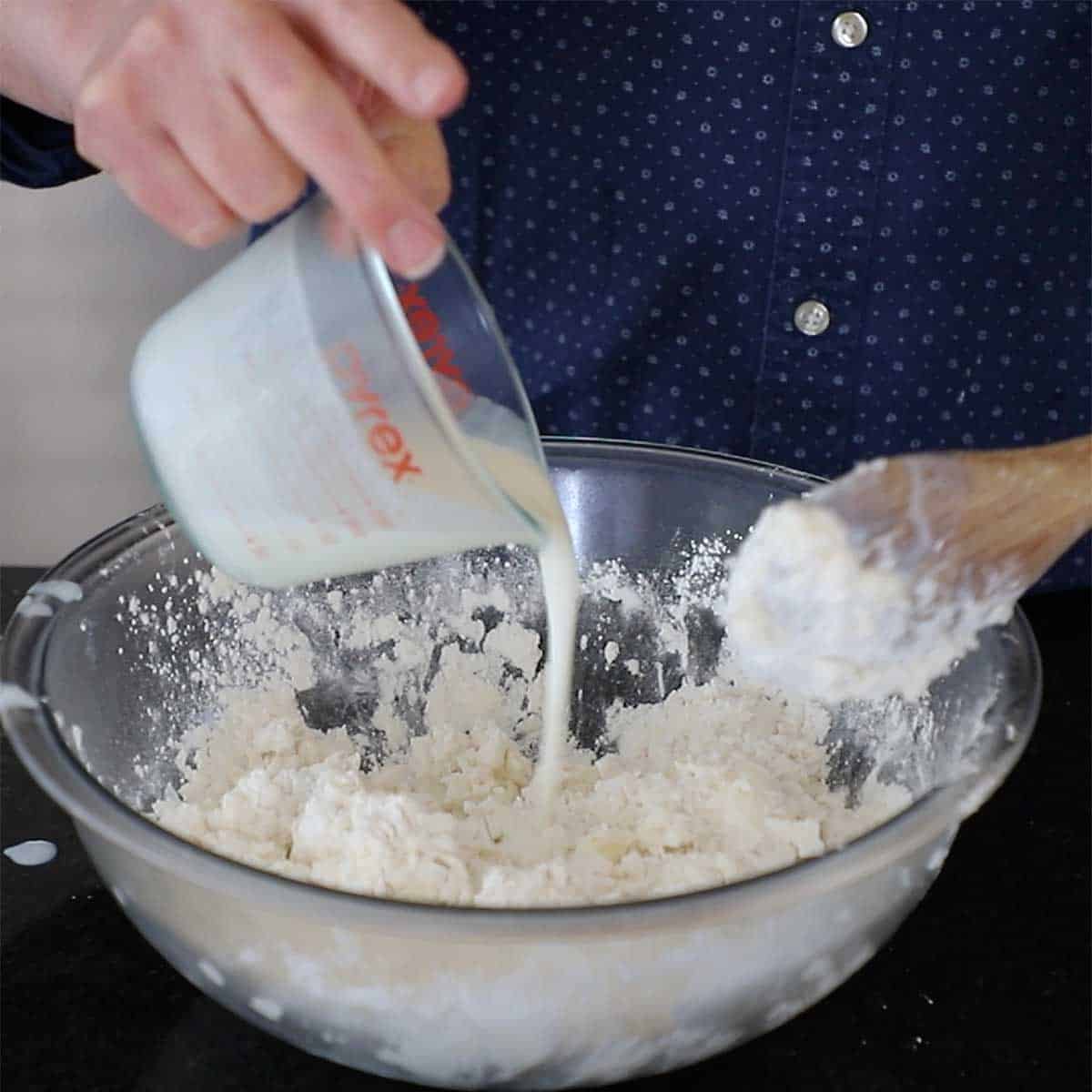A person pouring buttermilk from a glass measuring cup into a glass bowl filled with self-rising flour that has been mixed with chilled shredded butter.