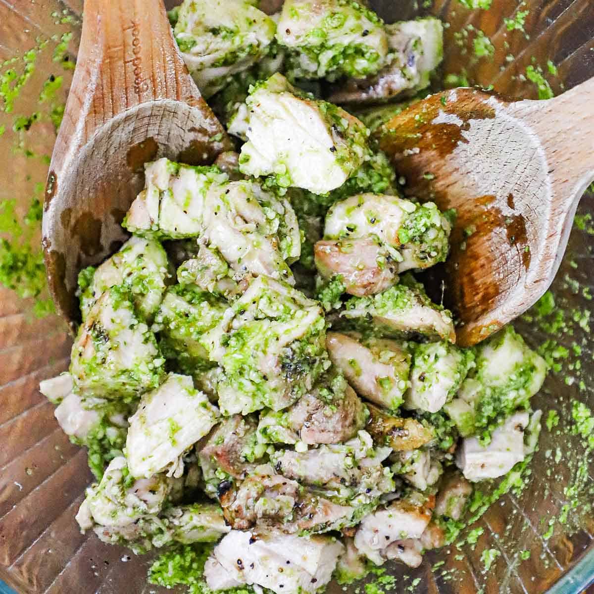 Grilled pieces of chicken that are being tossed by two wooden spoons in homemade pesto sauce in a large glass bowl.