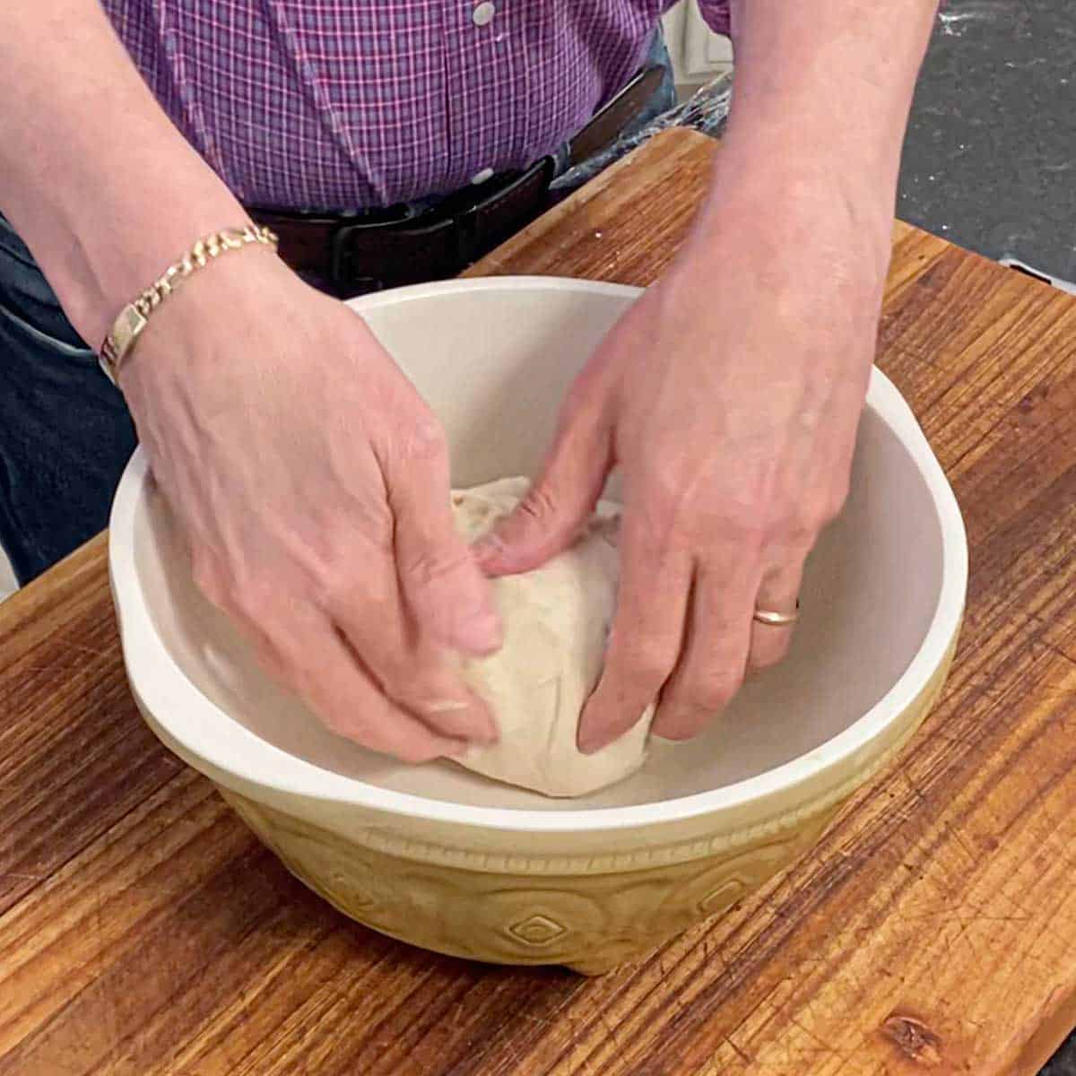 A person placing a ball of bagel dough into an oiled ceramic bowl sitting on a wooden cutting board.