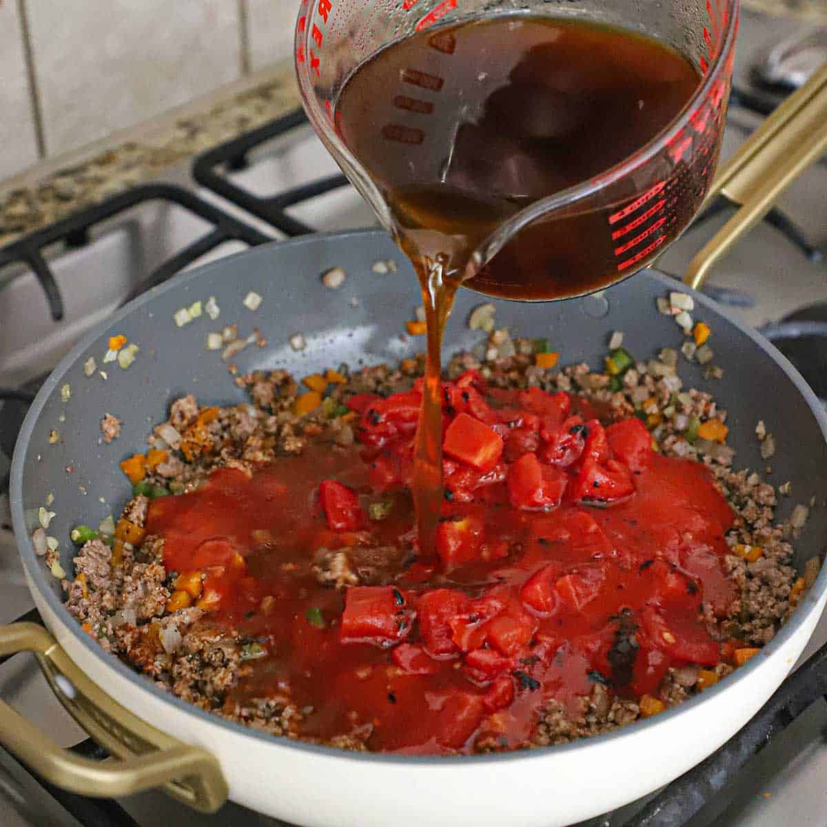A person pouring beef broth from a large glass measuring cup into a skillet filled with cooked ground beef, vegetables, chopped tomatoes, and tomato sauce.