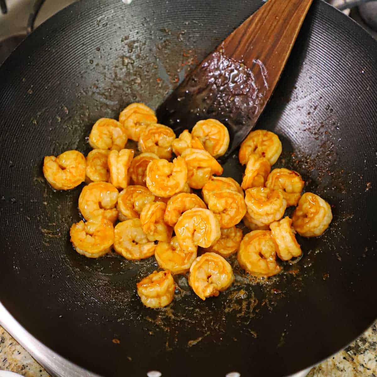 Marinated shrimp being sautéed in a large black wok with a wooden spatula off to the side.