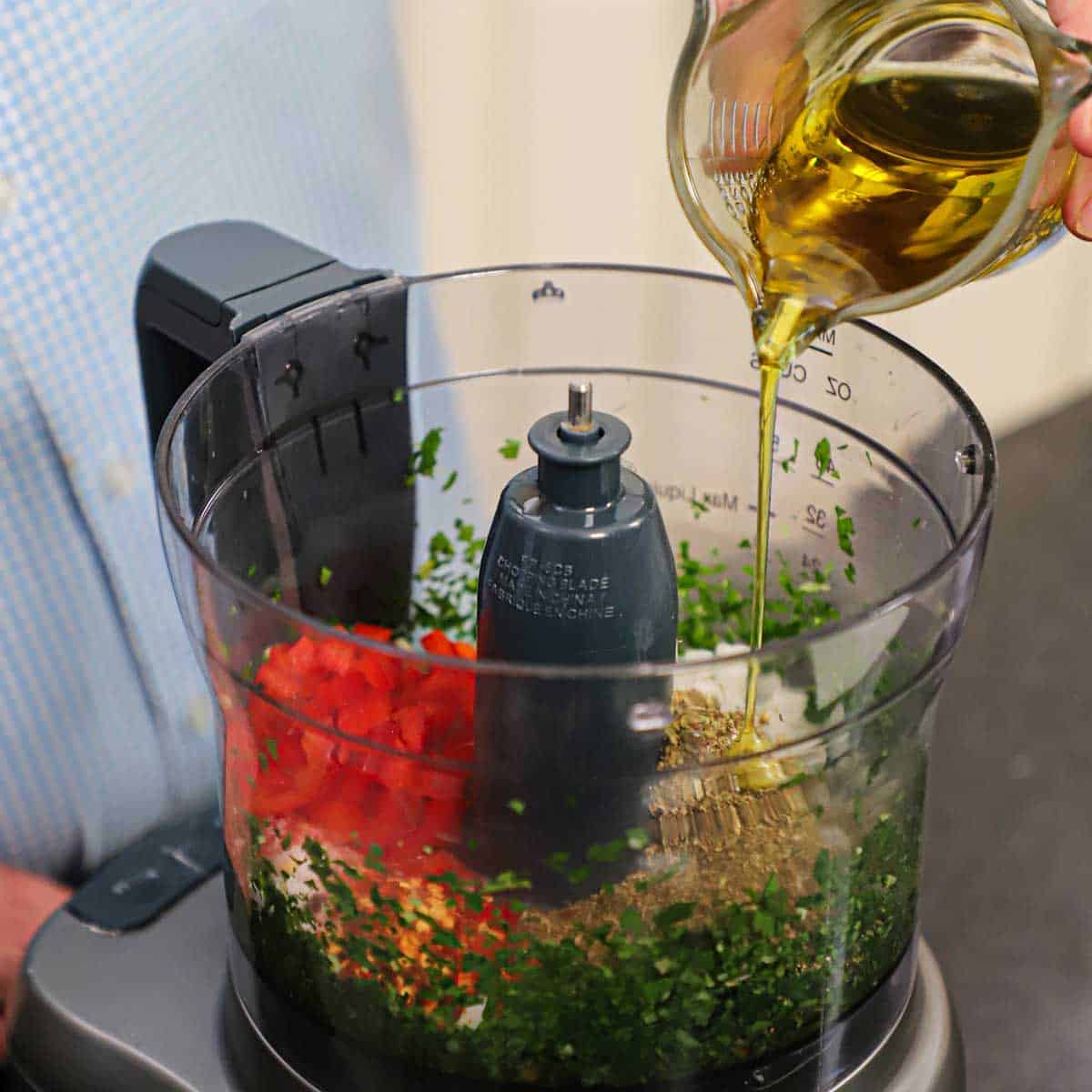 A person pouring extra-virgin olive-oil from a glass measuring cup into the base of a food processor that is filled with the ingredients for authentic chimichurri.